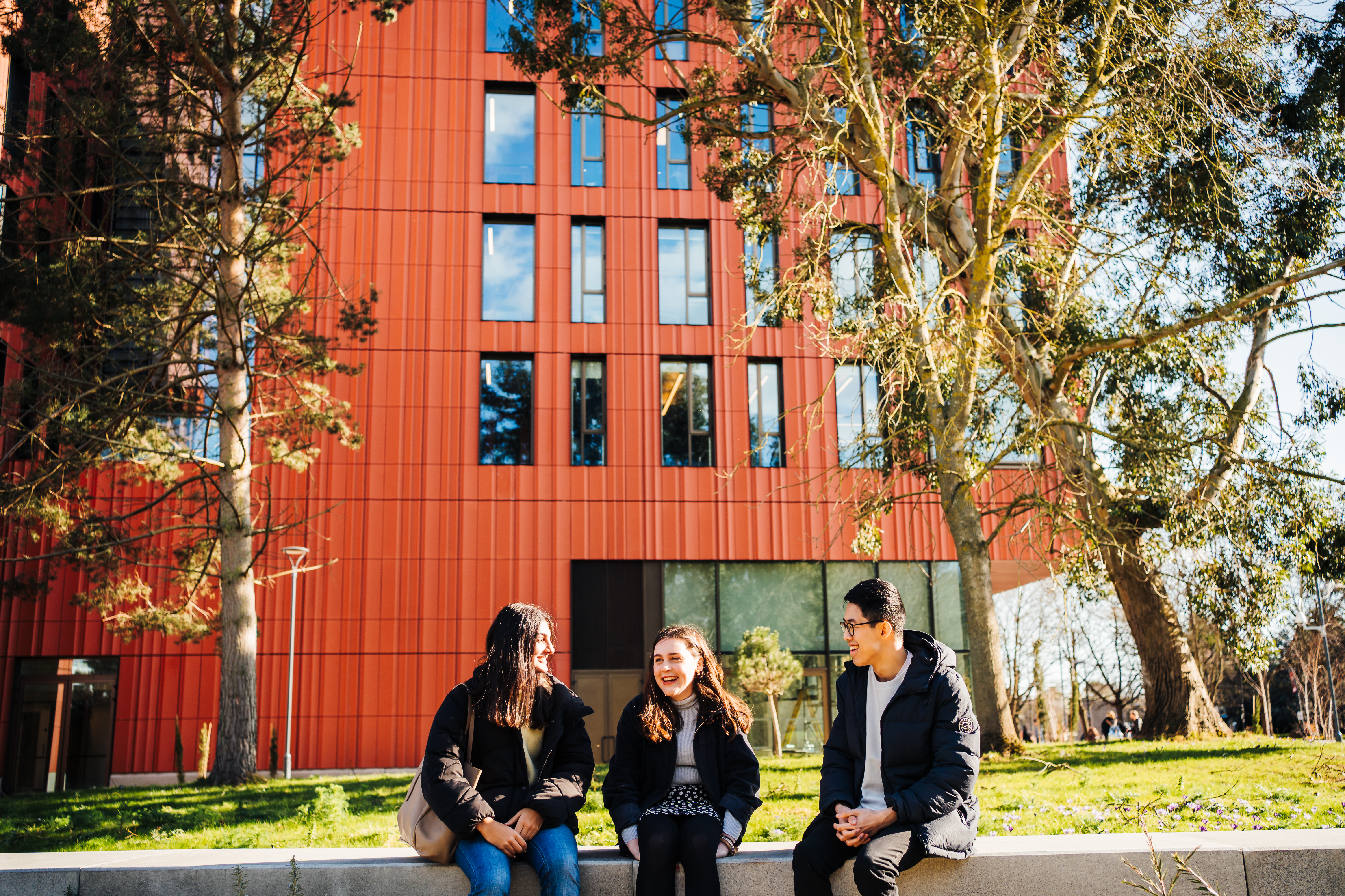 Three students laughing and chatting, sat near grass outside the faculty of art building
